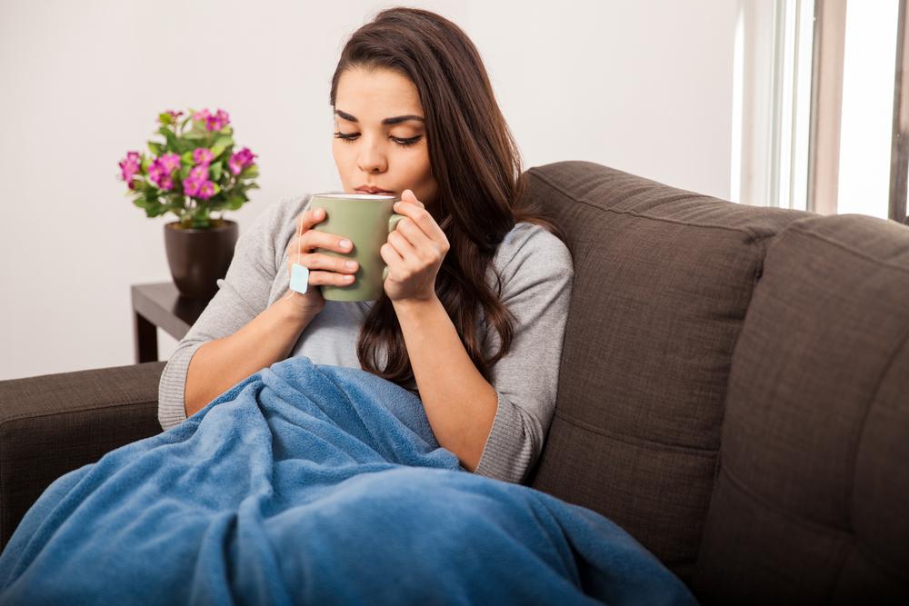 A woman bundled up on her couch drinking hot tea under a blanket.